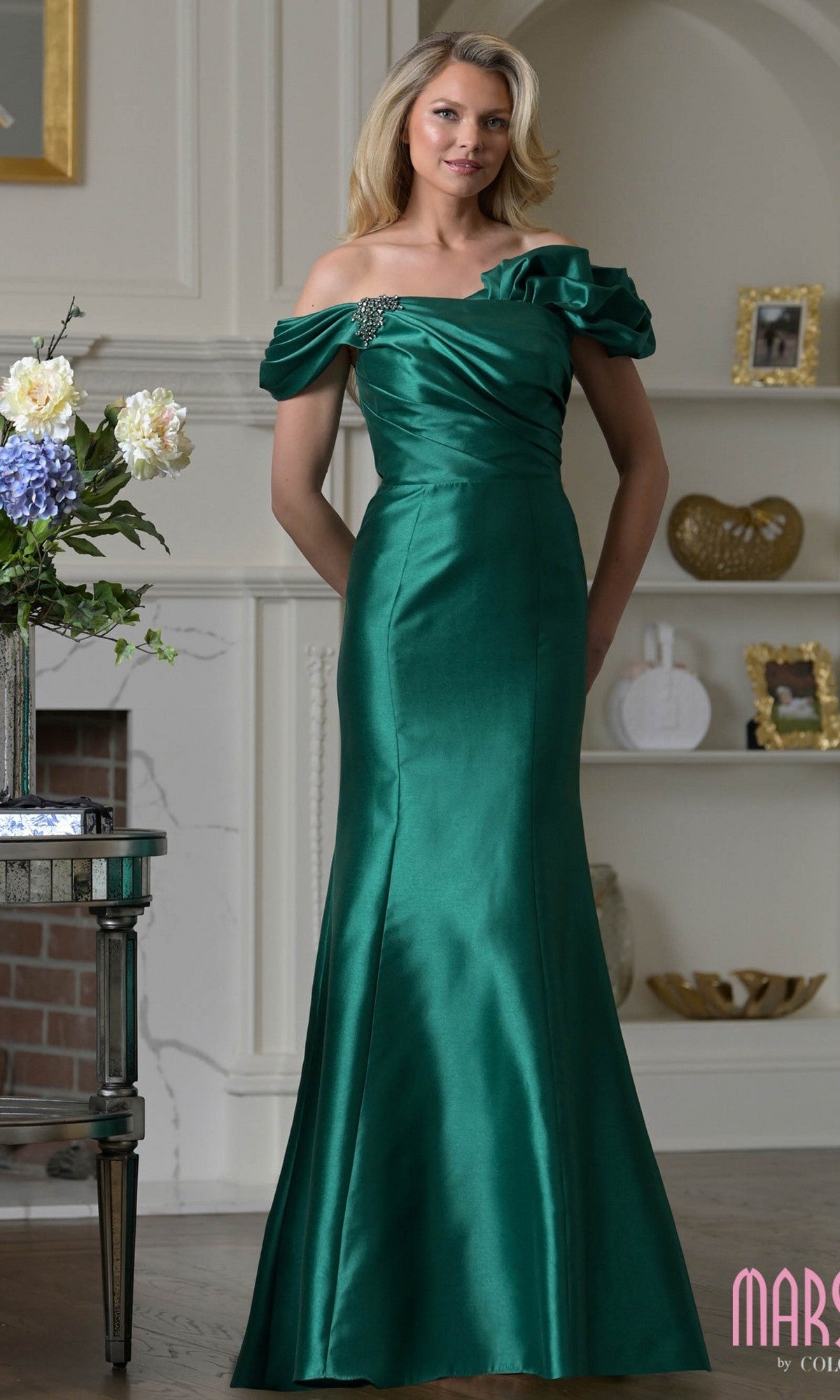 A woman wearing the Marsoni MV1407 long formal dress in emerald, featuring a ruffled off-shoulder design with a ruched bodice and floral accent, stands indoors near shelves and a fireplace.